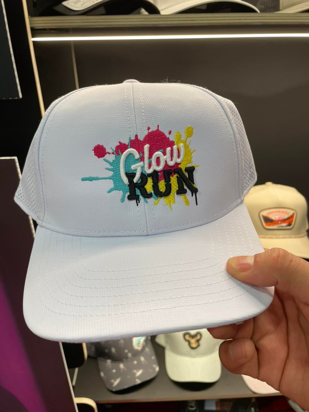 White trucker hat with a colorful embroidered Glow Run paint-splatter design