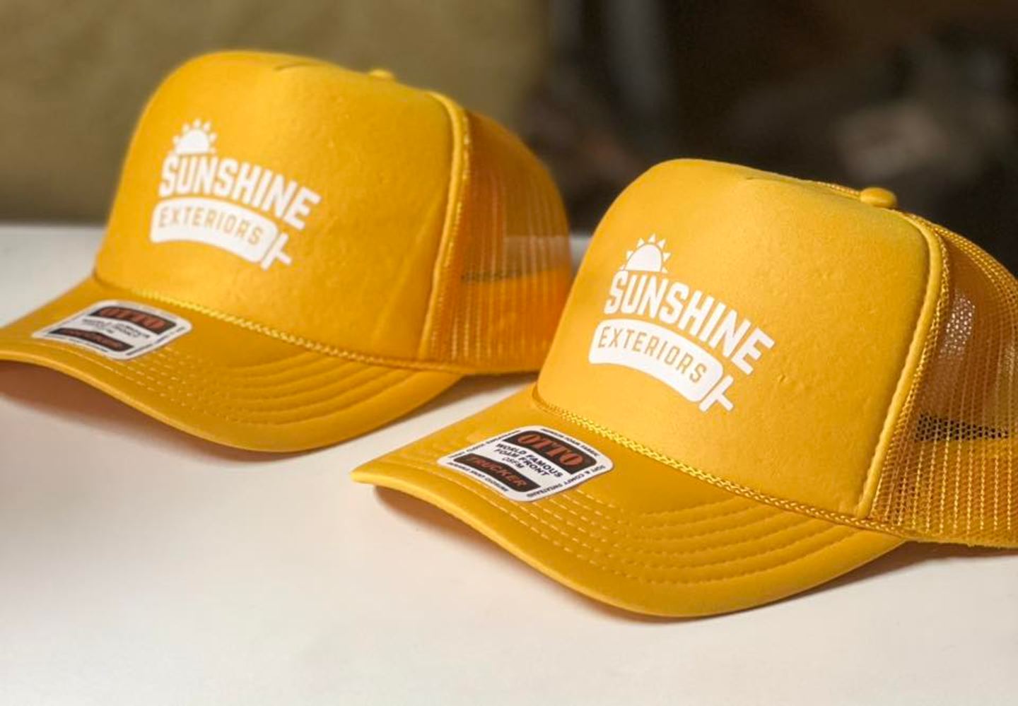 Two gold trucker hats with a white Sunshine Exteriors logo printed on the front panel